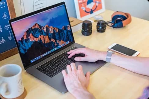 Person using a laptop on a wooden desk with an iPhone, headphones, camera lenses, and a coffee mug nearby